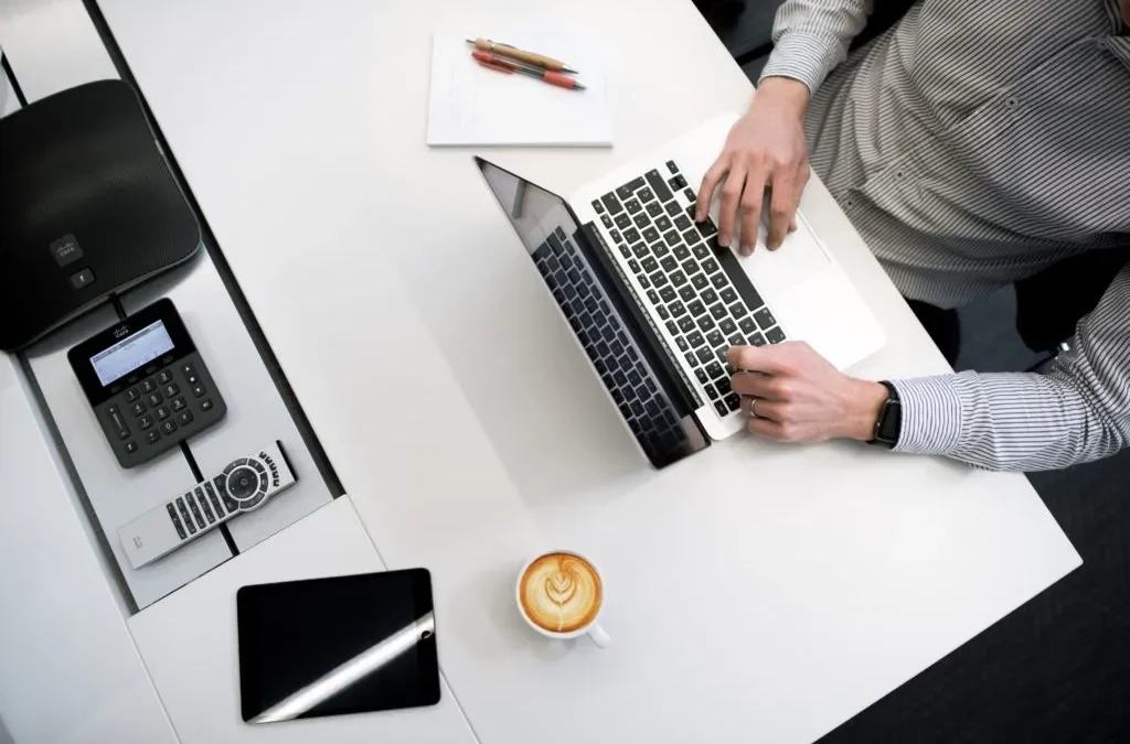 A person using a laptop on a white desk with tech accessories and a coffee cup.