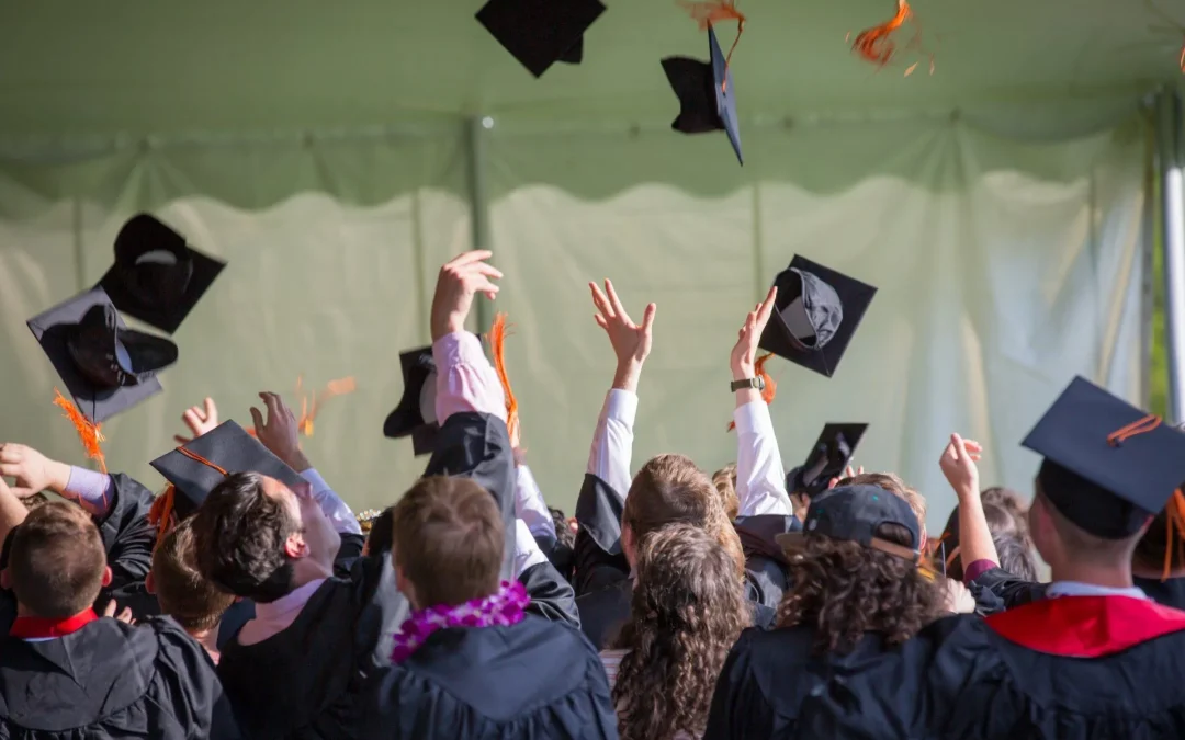Graduates tossing their caps in celebration, with a tent blurred in the background.