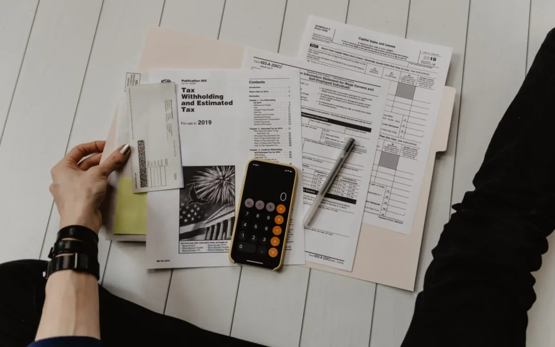 Overhead view of someone organizing tax forms with a calculator and pen on a white wooden surface.