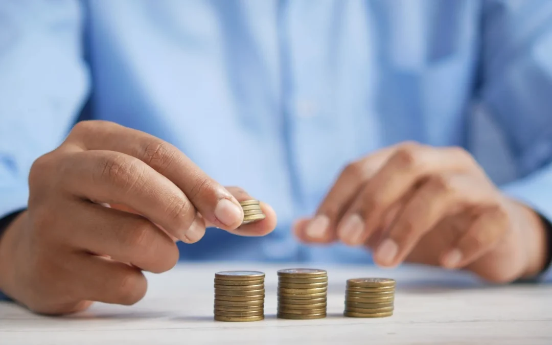 Person in a blue shirt is stacking coins in piles on a table, symbolizing money management or savings.