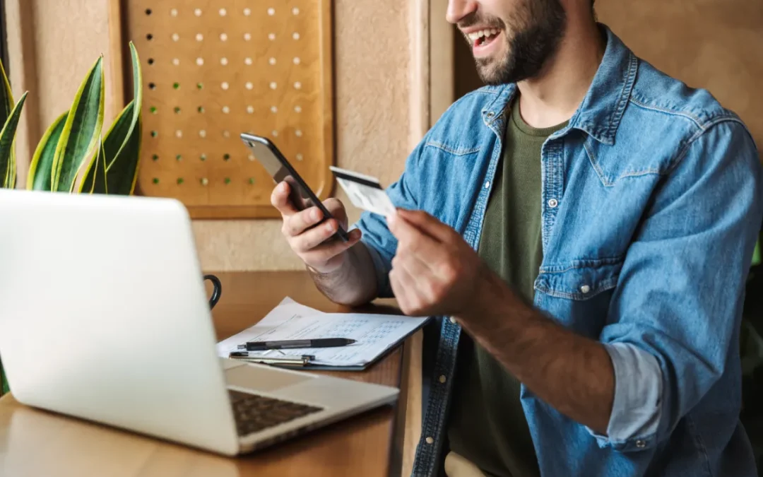 Smiling man using a smartphone and holding a credit card, with a laptop and papers on the desk
