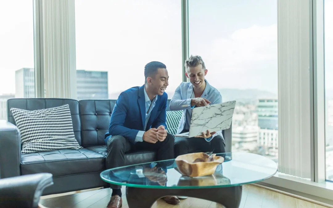 Two smiling men reviewing a laptop in a well-lit room with cityscape views.