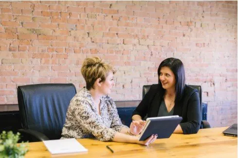 Two women, one with a tablet, are talking at a table in a brick-walled office.