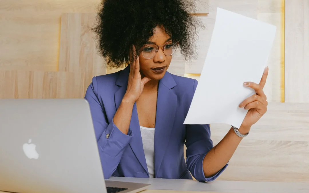 woman in a blue blazer reviews a document at her desk with a laptop open beside her.